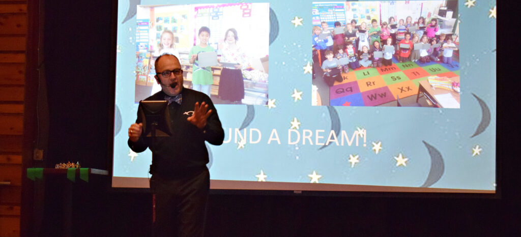 Presenter speaks on stage in front of classroom photos projection.