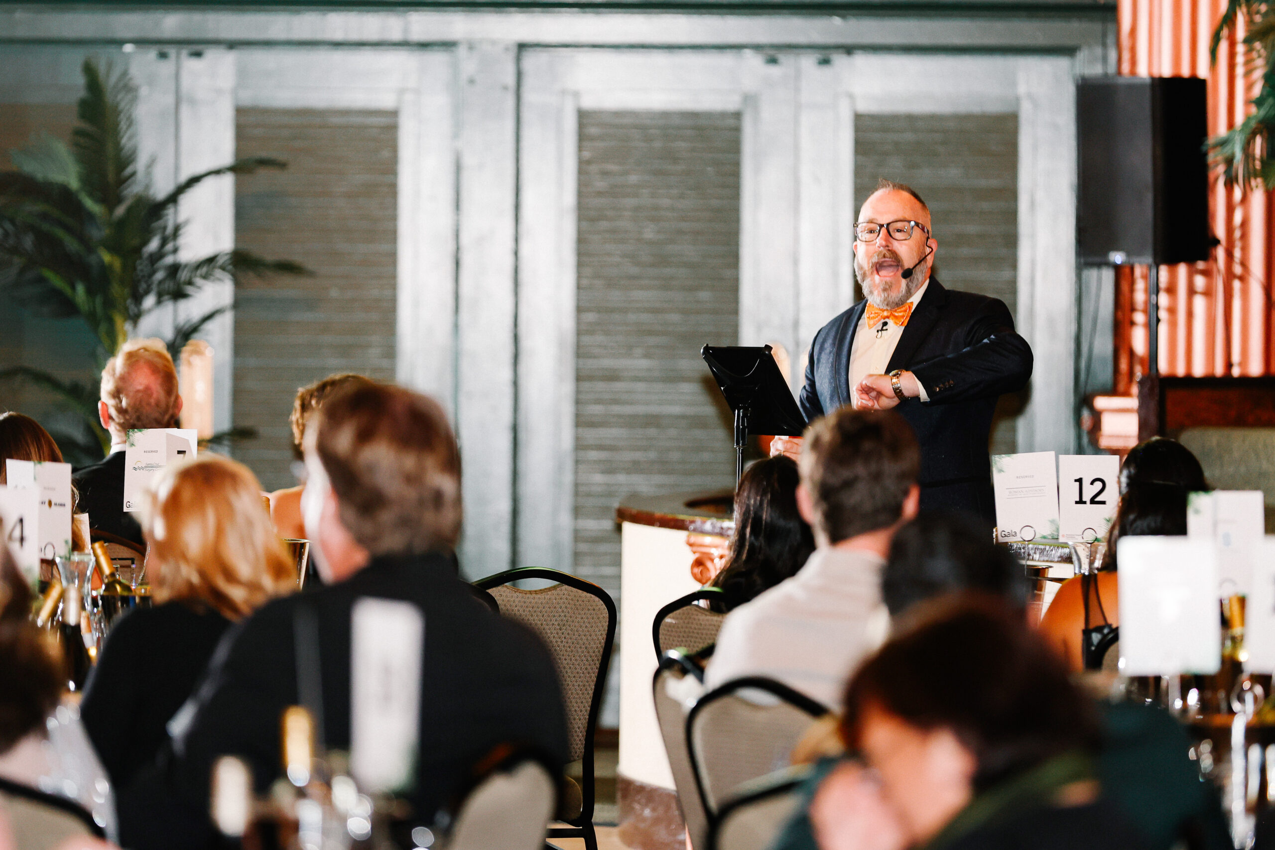 Host in dark jacket addresses engaged seated audience at indoor auction.