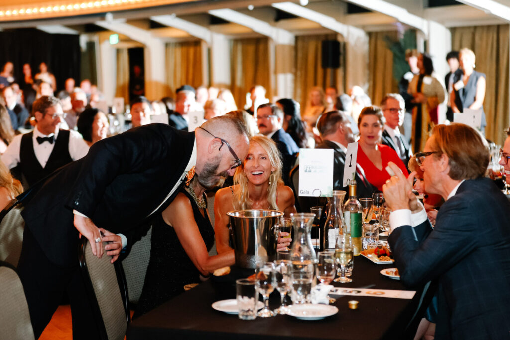 Host leans toward smiling guest during gala while others watch attentively.