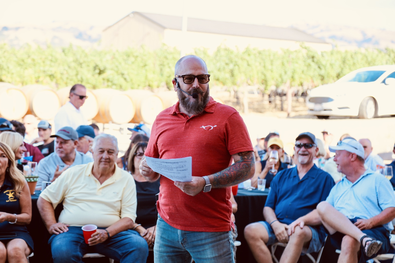 Auctioneer in red polo shirt engages cheerful outdoor crowd at winery.
