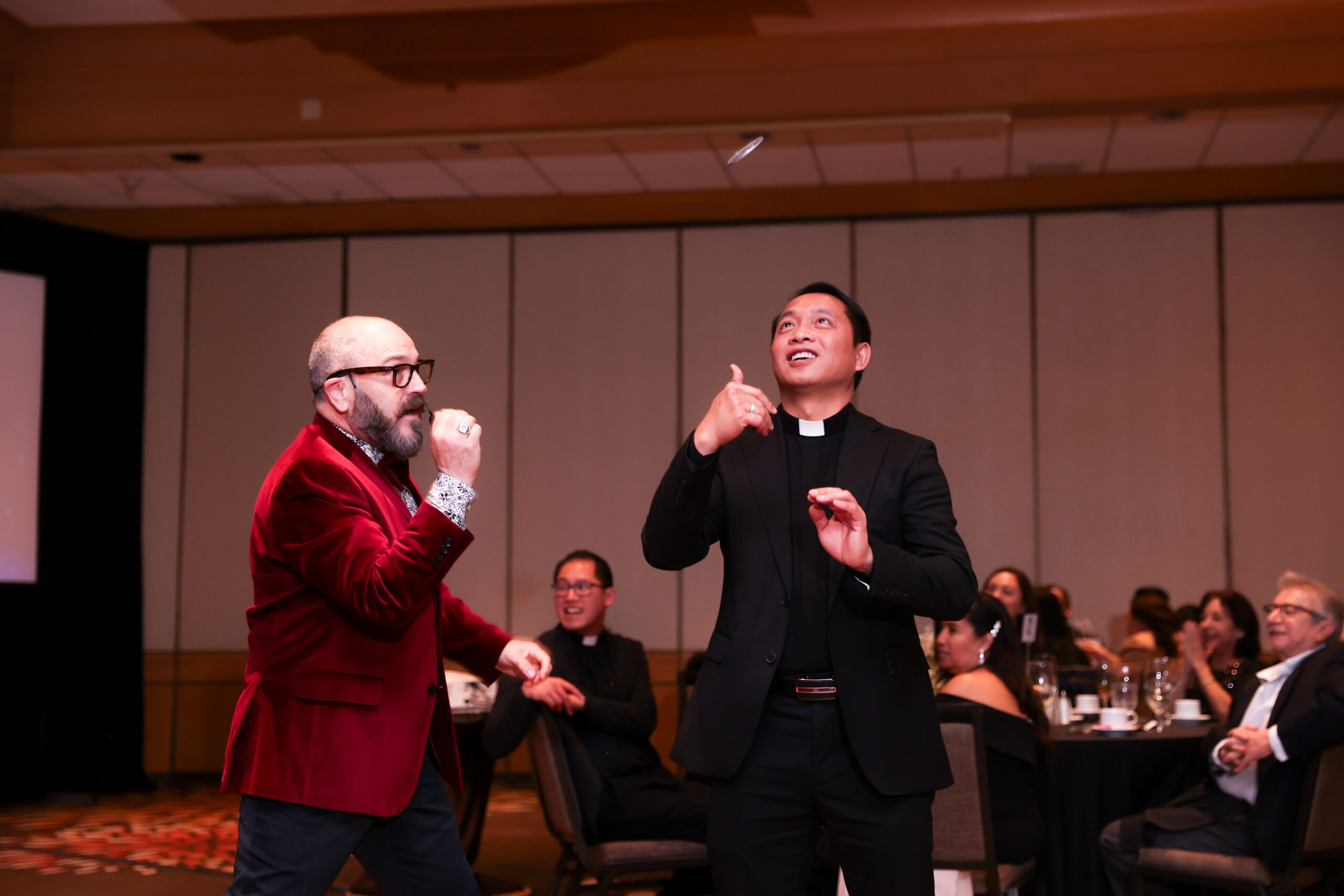 Host in red velvet jacket and priest laugh while tossing coin.