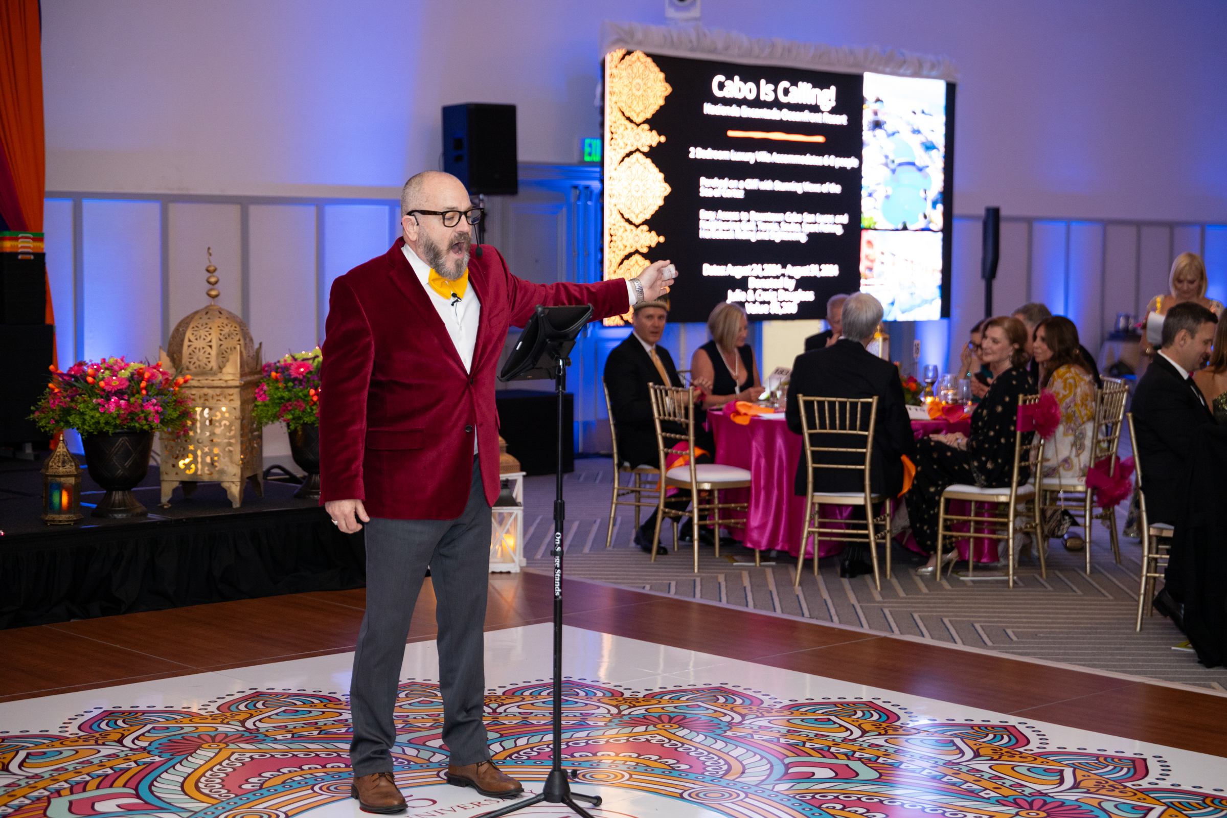 Auctioneer in red velvet blazer speaks on stage with event screen behind.
